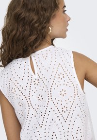 Blusa bianca ricamata con motivi a intaglio, caratterizzata da un dettaglio a chiave sul retro e design senza maniche, che mostra intricate motivazioni floreali.