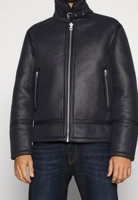 Svart läderjacka med hög krage, framdragkedja och sidofickor med dragkedjor. Strukturerad yta med horisontella detaljer i nederkanten.