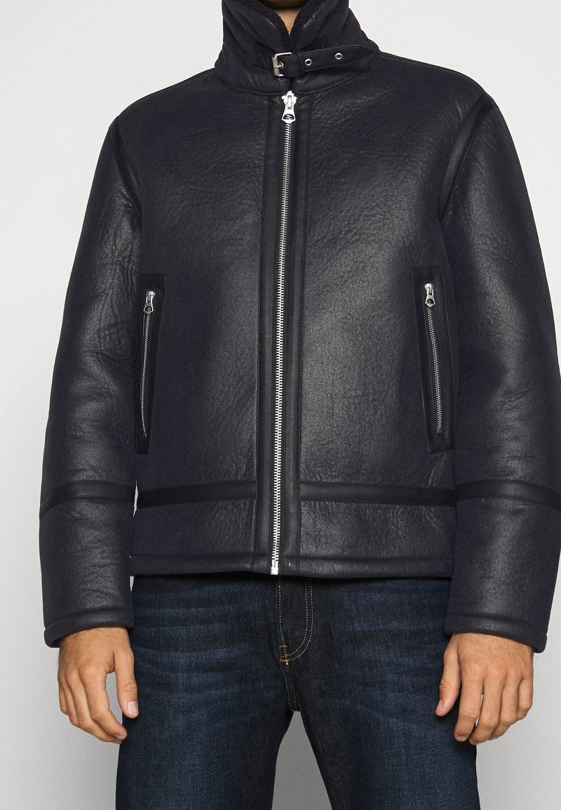 Svart läderjacka med hög krage, framdragkedja och sidofickor med dragkedjor. Strukturerad yta med horisontella detaljer i nederkanten.