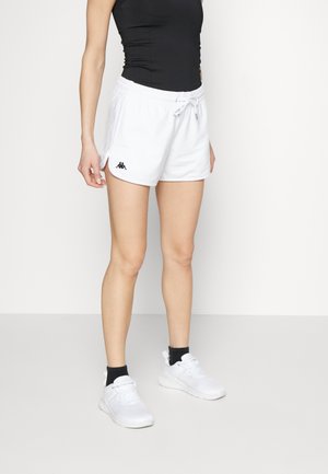 Persona con pantalones cortos deportivos blancos con cordón y logo negro, camiseta sin mangas negra, calcetines negros y zapatillas deportivas blancas, de pie sobre un fondo liso.