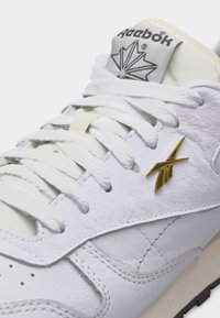 Κοντινή λήψη λευκού αθλητικού παπουτσιού Reebok με υφασμένο δέρμα, λευκά κορδόνια, χρυσό λογότυπο Reebok στο πλάι και ετικέτα στη γλώσσα με σχεδίαση φυλλομελά.