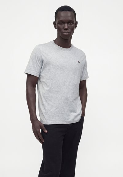 Homme à la peau foncée portant un t-shirt à manches courtes gris clair avec un petit logo brodé sur le côté gauche de la poitrine et un pantalon noir, debout devant un fond blanc.