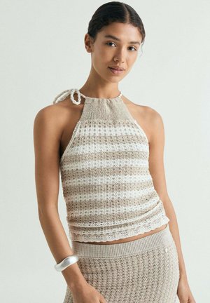 Mujer con top de punto sin mangas en beige y blanco a rayas con cuello halter y falda de punto texturizada a juego, con pulsera de plata en la muñeca.