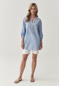 Tunica azzurra chiara a maniche lunghe con scollatura a V, due tasche frontali e bottoni. Abbinata a pantaloni corti bianchi e sandali chiari. Tessuto dalla consistenza liscia.