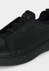 Baskets basses noires avec dessus synthétique texturé, bout rond, panneaux latéraux élastiques et semelle en caoutchouc noire sur fond blanc.