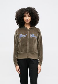 Jovem com cabelo encaracolado vestindo um casaco com capuz castanho com fecho e letras azuis "Joop! Jeans" e calças pretas, em pé contra um fundo branco.