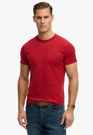 Homme portant un t-shirt rouge à manches courtes, un jean bleu avec ceinture marron et une montre-bracelet, debout devant un fond blanc.