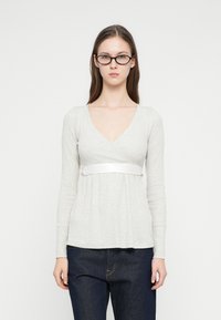 Γυναίκα με μακριά καστανά μαλλιά που φοράει μαύρα γυαλιά, γκρι ανοιχτόχρωμο μπλουζάκι με μακριά μανίκια και V λαιμόκοψη με λευκή ταινία, και σκούρο μπλε τζιν, στέκεται μπροστά σε λευκό φόντο.