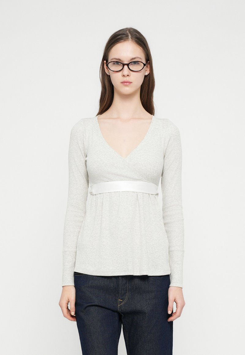 Γυναίκα με μακριά καστανά μαλλιά που φοράει μαύρα γυαλιά, γκρι ανοιχτόχρωμο μπλουζάκι με μακριά μανίκια και V λαιμόκοψη με λευκή ταινία, και σκούρο μπλε τζιν, στέκεται μπροστά σε λευκό φόντο.