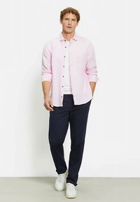 Hellrosa Hemd mit Knöpfen und hochgekrempelten Ärmeln, weißes T-Shirt und marineblaue Hose. Das Modell trägt weiße Sneaker und steht vor einem weißen Hintergrund.