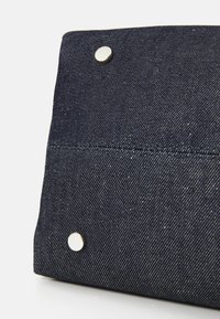 Denim-laukku tummansinisellä värillä, jossa on sileä pinta, litteä muotoilu ja edessä kaksi metallista nappia.