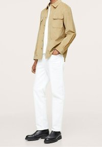 Mand iført en beige skjorte med knapper, hvide bukser og sorte klodsede sko, der står med den ene hånd i lommen mod en ensfarvet baggrund.