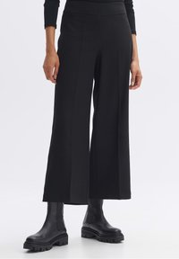 Pantalon noir à jambes larges en tissu lisse avec une taille haute, associé à des bottines noires épaisses. Détails de coutures visibles et texture.