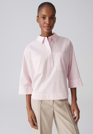 Vrouw draagt een lichtroze, loszittend overhemd met een kraag en twee borstzakken, en beige broek, staand tegen een effen lichte achtergrond.