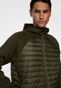 Man i olivgrön quiltad jacka med dragkedja framtill och huva, som tittar åt sidan mot en neutral vit bakgrund.