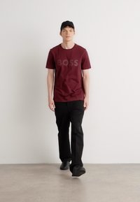 Burgundy tričko s logom "BOSS", skombinované s čiernymi nohavicami a čiernymi teniskami. Model v čiernej šiltovke, stojaci na jednoduchom pozadí.