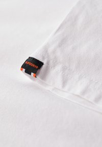 Biały bawełniany materiał o prążkowanej fakturze, z czarną metką z napisem "SUPERDRY" w kolorze pomarańczowym oraz dwoma pomarańczowo-białymi akcentami w pobliżu szwu.