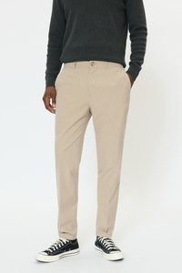 Beżowe spodnie o dopasowanym kroju, z gładkim przodem, bocznymi kieszeniami i zapięciem na guzik. Noszone z czarnymi sneakersami.