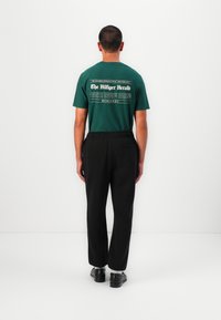 Grünes Baumwoll-T-Shirt mit weißem Druck auf dem Rücken. Schwarze Hosen mit elastischem Bund, kombiniert mit dunklen Schuhen.