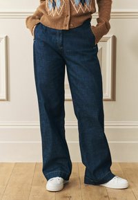 Plati kāju tumši zila džinsu bikses ar priekšējiem kabatām, apvienotas ar baltām trenerēm. Audumam ir smalka tekstūra un kontrasta šuvju detaļas.