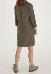 Robe chemise vert olive avec un col, manches 3/4, coupe droite et texture douce. Associée à des bottines gris clair.