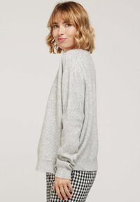 Pull gris clair présenté, avec une coupe décontractée, des poignets côtelés et un col rond, porté sur un pantalon à carreaux noir et blanc.