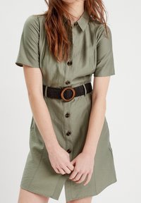 Robe chemise vert olive à manches courtes, devant à boutons et poches latérales. Ceinture tissée noire avec une boucle en bois circulaire.