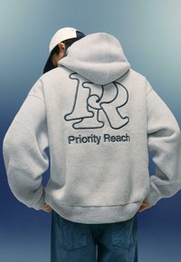 Svetlo šedá mikina s textúrovaným vzorom, na chrbte s veľkým tmavomodrým vyšitým "1R" a "Priority Reach". Kapucňa a vrecko sú viditeľné.