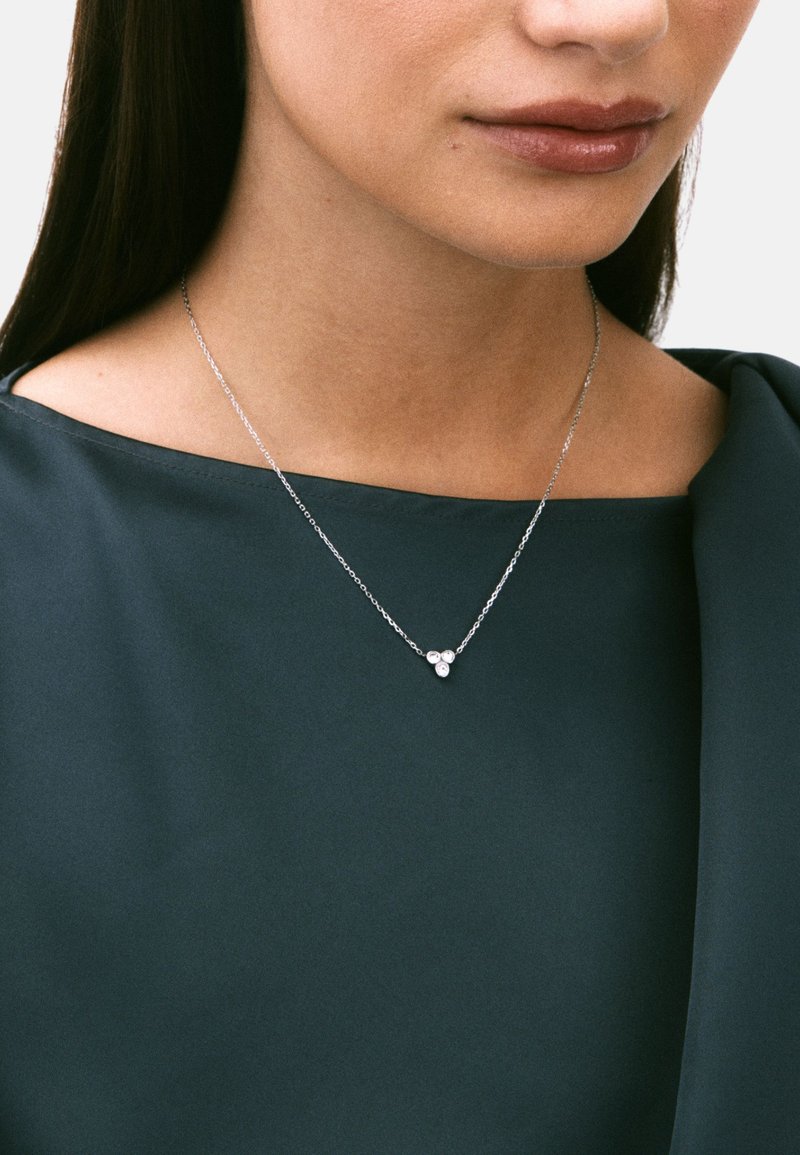 Sølvhalskæde med tre cirkulære vedhæng, båret til en mørkegrøn top med glat tekstur og bred udskæring.