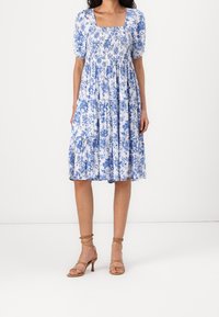 Robe fleurie bleu et blanche avec un décolleté carré, des manches bouffantes et une jupe à volants ; complétée par des talons beiges à brides.