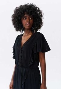 Robe noire avec des manchettes à volants, décolleté en V avec bordure festonnée et taille marquée. Tissu lisse avec un éclat subtil et silhouette fluide.