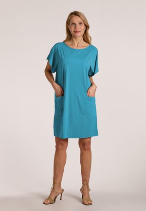 Kvinde iført løs kortærmet teal kjole med store forlommer, stående med hænderne i lommerne, beige højhælede sandaler.