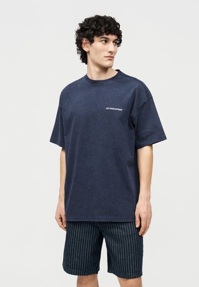 Jeune homme aux cheveux bouclés portant un t-shirt surdimensionné bleu marine avec le texte "LES BENJAMINS" et un short à fines rayures foncées, regardant de côté.