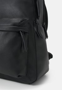 Sac à dos noir en simili cuir avec une texture lisse, doté de poches zippées et de tirettes assorties. Design arrondi et coutures renforcées.