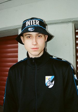 Zwarte zip-up jas met blauwe accenten en logo; bijpassende zwarte bucket hat met de tekst "INTER". Haar is kort en heeft een casual stijl.