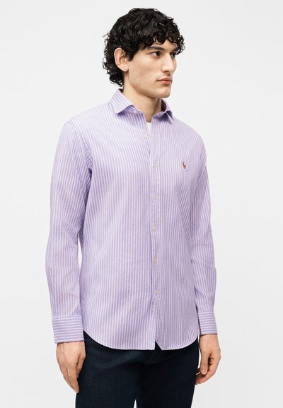 Άνδρας που φοράει μακρυμάνικο πουκάμισο με μοβ και λευκές ρίγες με κουμπιά, στέκεται με τα χέρια χαλαρά, κοιτάζοντας προς την πλευρά.