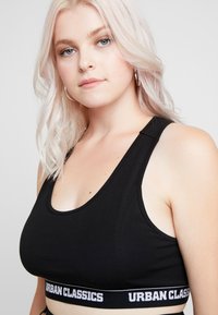 Svart sportbehå med scoop-urringning, breda axelband och ett märkt elastiskt band med "URBAN CLASSICS" i vita bokstäver.