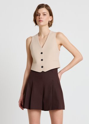 Giovane donna con capelli corti che indossa un gilet beige senza maniche con bottoni neri e una gonna plissettata marrone scuro.
