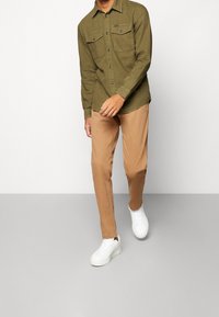 Oliivinvihreä pitkähihainen paita, jossa kaksi rintataskua, vaaleanruskeat housut ja valkoiset lenkkarit. Sileä pinta, napillinen malli ja rento istuvuus.