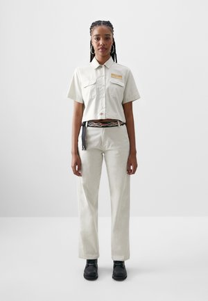 Camisa beige corta con mangas cortas y dos bolsillos en el pecho, combinada con pantalones de tiro alto beige claro y zapatos negros.