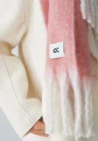 Sciarpa in gradiente rosa e bianco con estremità frangiate, realizzata in materiale morbido. Include un piccolo etichetta bianca con un logo nero.