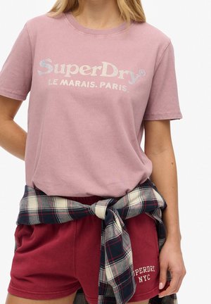 Kobieta w pudroworóżowej koszulce SuperDry z napisem "Le Marais. Paris.", bordowych szortach i kraciastej koszuli związanej wokół talii.