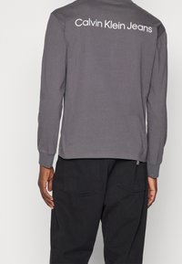 Harmaa puuvillapaita pitkillä hihoilla, jossa on pyöreä pääntie ja valkoinen "Calvin Klein Jeans" -logo selässä, yhdistettynä tummiin housuihin.