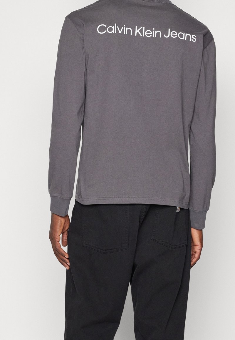 Grijs katoenen t-shirt met lange mouwen, voorzien van een ronde hals en een wit "Calvin Klein Jeans" logo op de achterkant, gecombineerd met donkere pants.