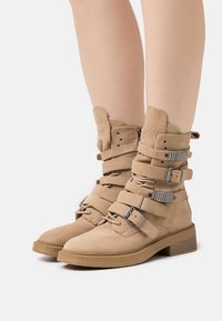 Botins de tornozelo em camurça taupe com múltiplas tiras ajustáveis, detalhes em metal prateado e sola de borracha texturizada. Bico quadrado e tacão baixo.