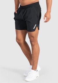 Pantalones cortos deportivos negros con una cintura elástica, bolsillos laterales y un dobladillo en diagonal. Presenta una marca en blanco en la pierna izquierda.