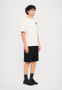 Short-sleeve cream cotton t-shirt with a small black graphic, paired with black shorts. Model wears dark socks and black loafers.