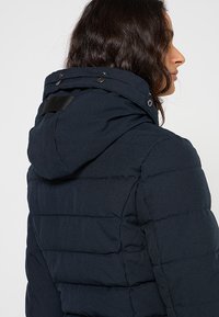 Giacca imbottita blu navy con cappuccio, caratterizzata da un design trapuntato, bottoni a pressione sul colletto e tessuto strutturato. Vista posteriore mostra una forma aderente.