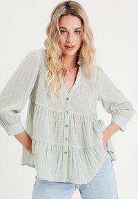 Blouse rayée verte et blanche avec un design à volants, fermeture à boutons sur le devant et une poche poitrine unique. Manches longues avec des poignets.