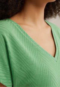 Detailní záběr osoby v zeleném texturovaném topu s V-výstřihem a šikmým vzorem, zobrazuje oblast krku a ramen.